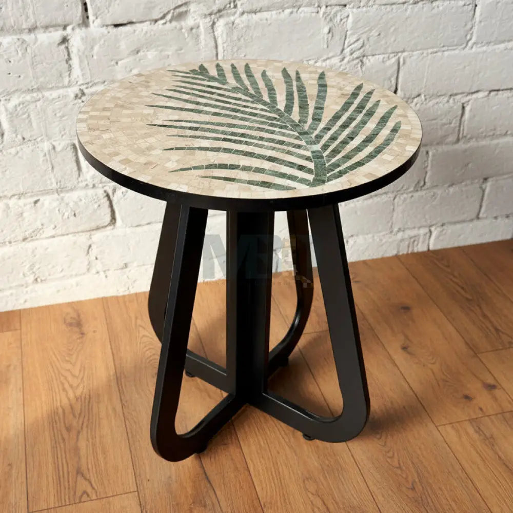 A round coffee table with a nature-inspired stone top featuring a tea leaf pattern, supported by a bold black infinity loop base, placed in a room with a white brick wall and patterned floor tiles.