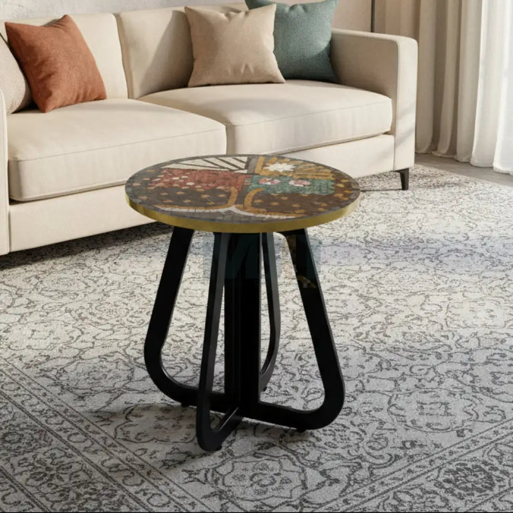 A round mosaic coffee table with a tree-inspired pattern on the tabletop and an ornate curved base, placed on a tiled floor against a white brick wall background with a potted plant and decorative tiles.