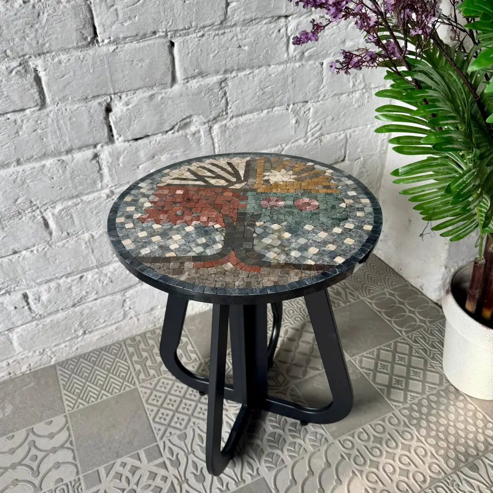 A round coffee table with a tree of life design and an infinity loop base, placed on a tiled floor next to a potted plant.